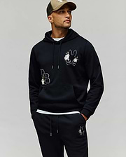 A man wears the Psycho Bunny MENS AUGUSTE HOODIE - MT0500043 in black with cartoon bunny patches and chenille floral details, matching sweatpants, and a beige cap, standing hands-in-pockets against a plain background.