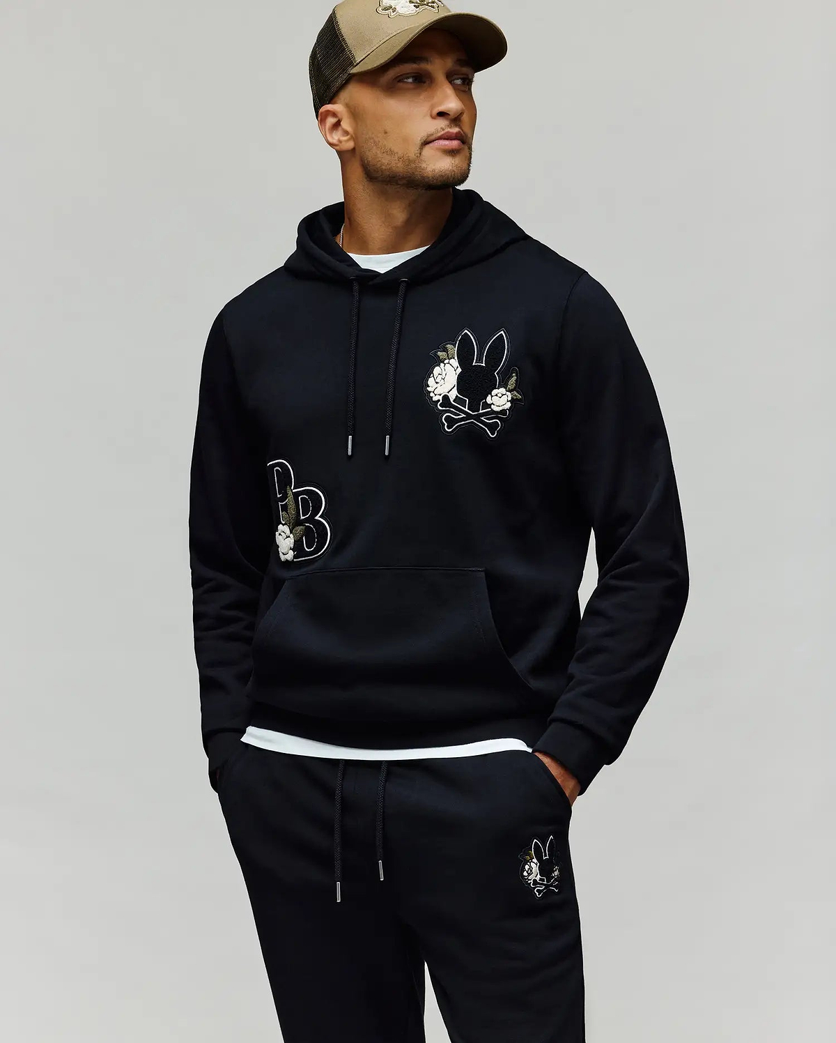 A man wears the Psycho Bunny MENS AUGUSTE HOODIE - MT0500043 in black with cartoon bunny patches and chenille floral details, matching sweatpants, and a beige cap, standing hands-in-pockets against a plain background.
