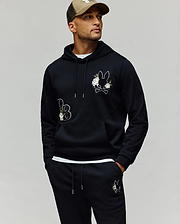 A man wears the Psycho Bunny MENS AUGUSTE HOODIE - MT0500043 in black with cartoon bunny patches and chenille floral details, matching sweatpants, and a beige cap, standing hands-in-pockets against a plain background.