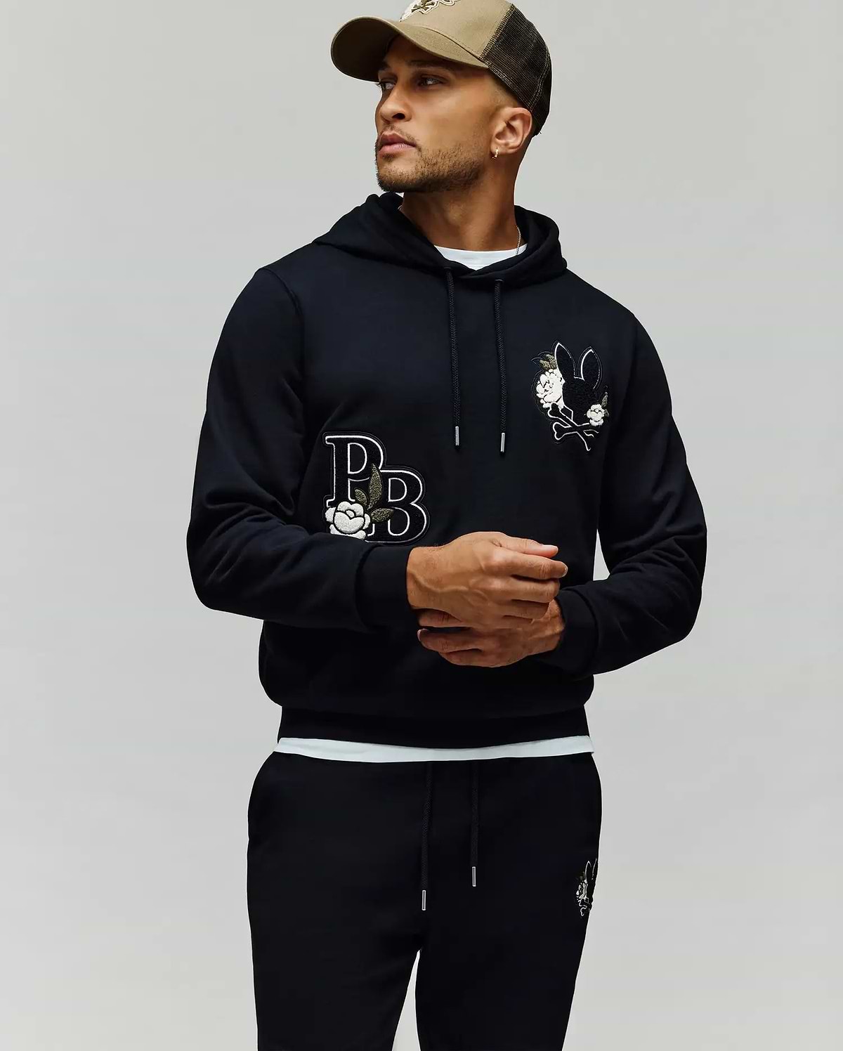 A man in the Psycho Bunny MENS AUGUSTE HOODIE (MT0500043) with cartoon graphics, chenille floral details, and "PB" lettering pairs matching sweatpants and a brown cap, standing against a plain light background, looking to the side.