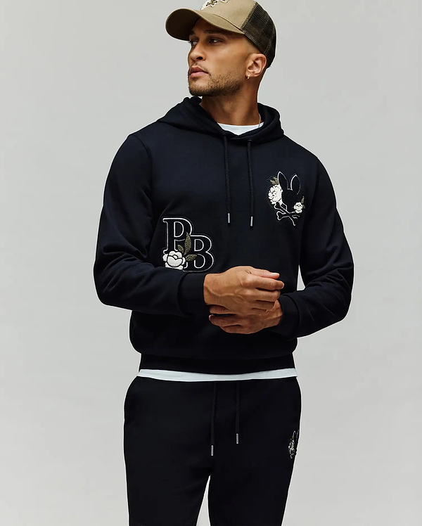 A man in the Psycho Bunny MENS AUGUSTE HOODIE (MT0500043) with cartoon graphics, chenille floral details, and "PB" lettering pairs matching sweatpants and a brown cap, standing against a plain light background, looking to the side.