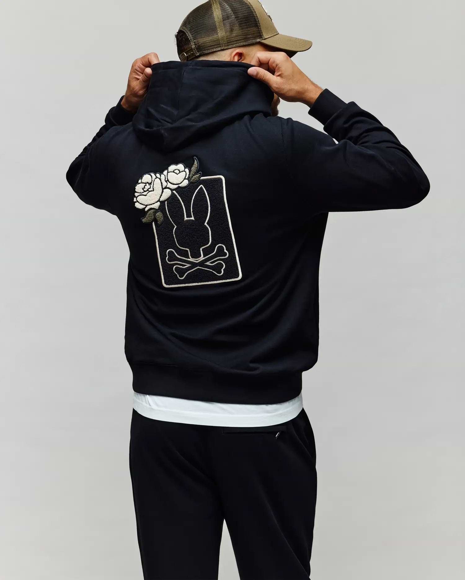 A person wears the Psycho Bunny MENS AUGUSTE HOODIE (MT0500043), a regular fit premium cotton hoodie with chenille floral detailing, bunny, crossbones, and white roses on the back, adjusting a brown-tan cap while facing away.