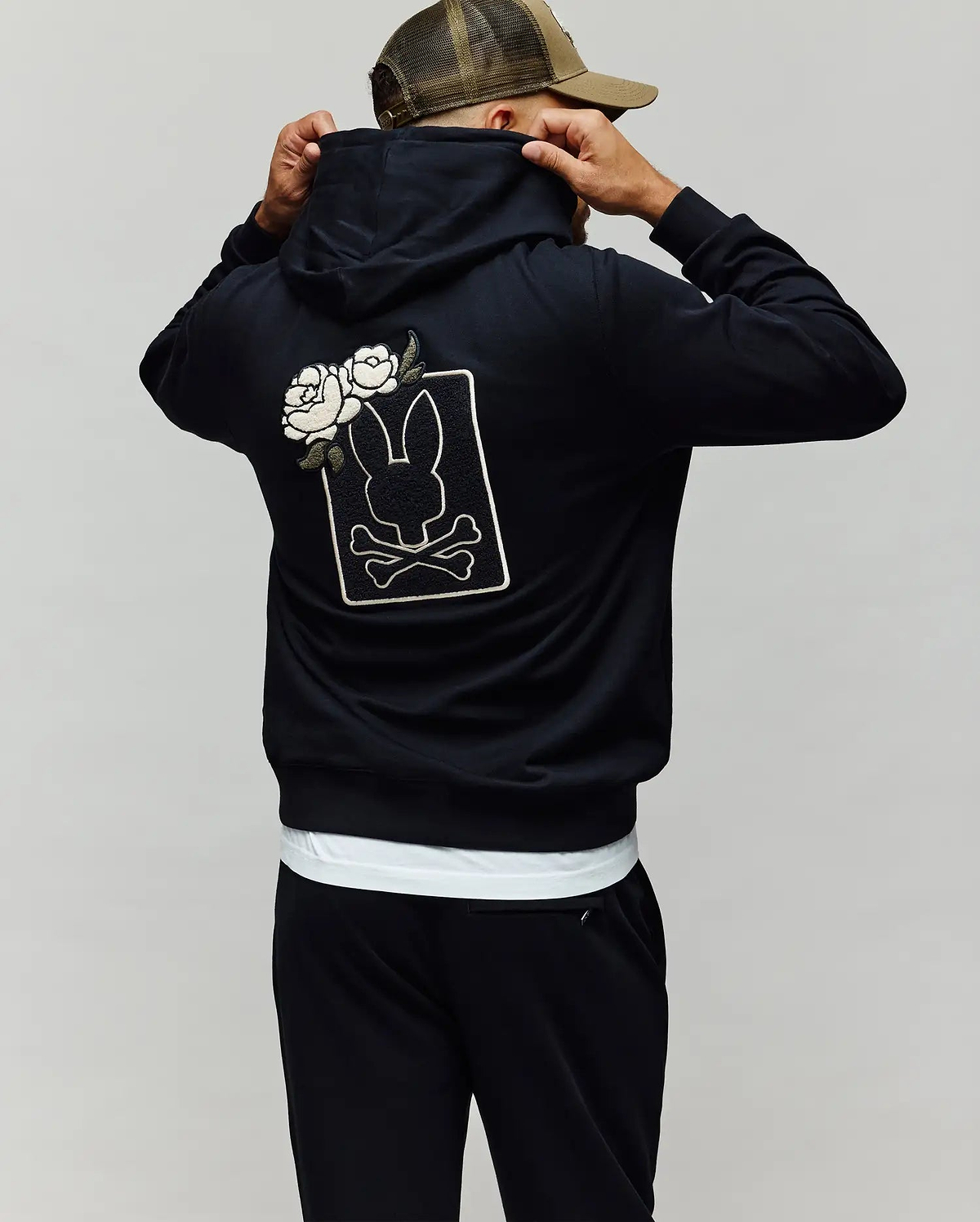 A person wears the Psycho Bunny MENS AUGUSTE HOODIE (MT0500043), a regular fit premium cotton hoodie with chenille floral detailing, bunny, crossbones, and white roses on the back, adjusting a brown-tan cap while facing away.