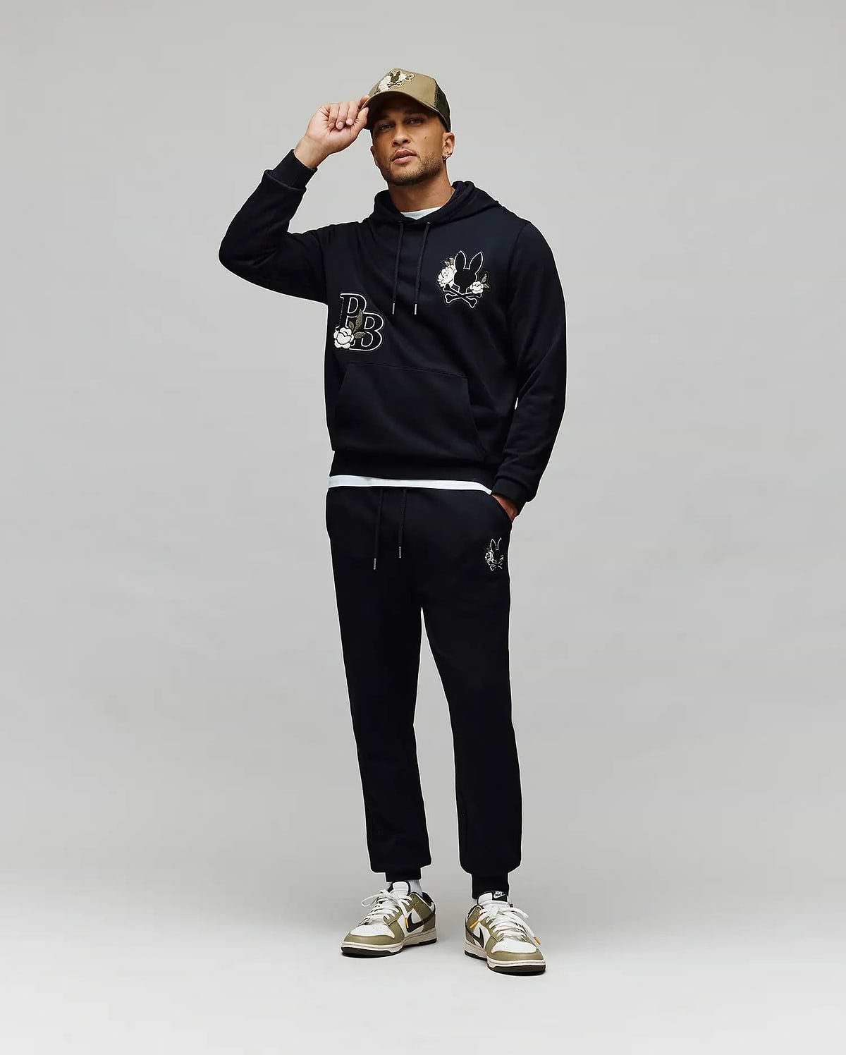 A man poses against a gray background, touching the brim of his beige cap while wearing Psycho Bunny’s MENS AUGUSTE HOODIE (MT0500043), matching joggers, and sneakers. The hoodie features regular fit premium cotton and chenille floral detailing.