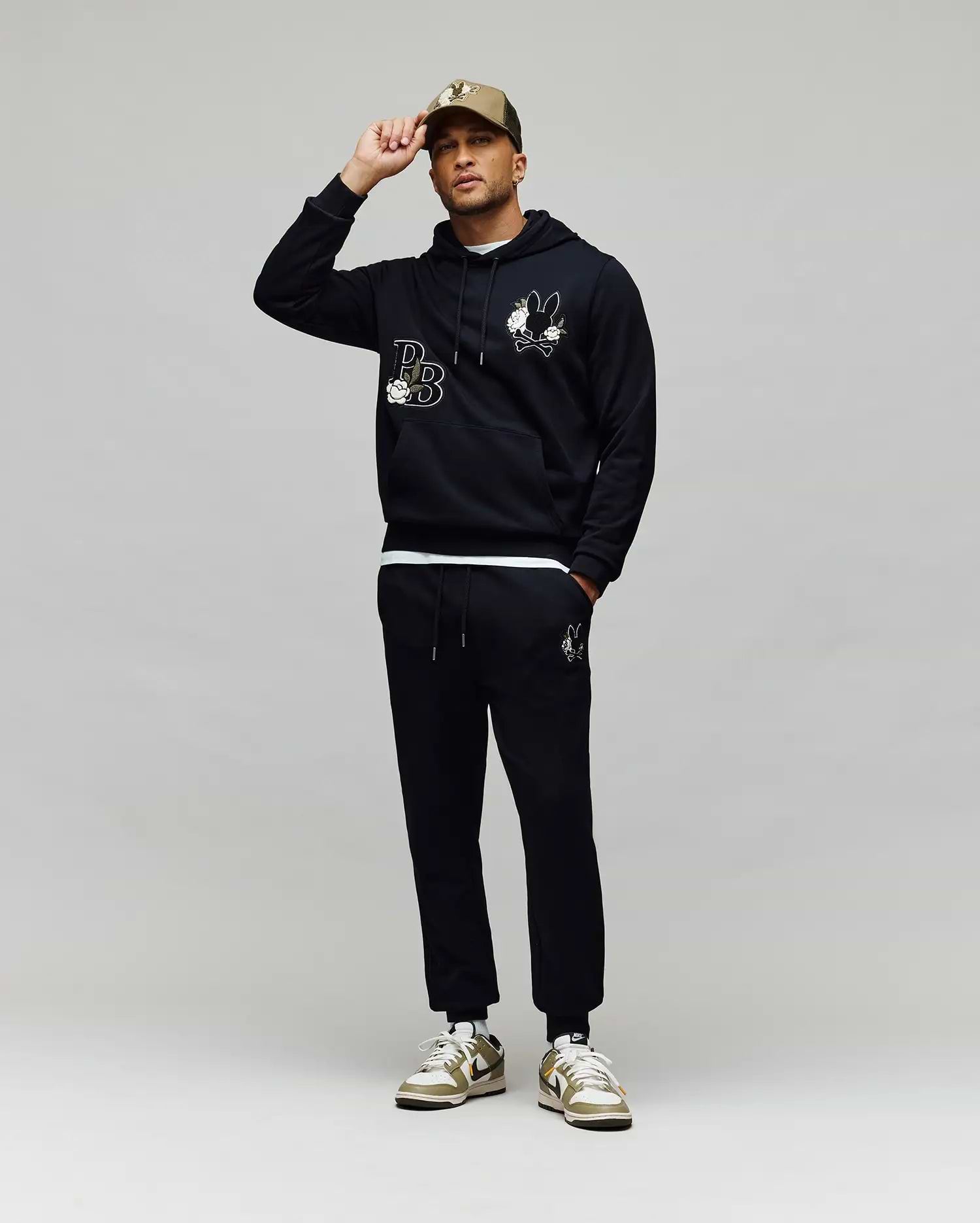 A man poses against a gray background, touching the brim of his beige cap while wearing Psycho Bunny’s MENS AUGUSTE HOODIE (MT0500043), matching joggers, and sneakers. The hoodie features regular fit premium cotton and chenille floral detailing.