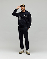A man poses against a gray background, touching the brim of his beige cap while wearing Psycho Bunny’s MENS AUGUSTE HOODIE (MT0500043), matching joggers, and sneakers. The hoodie features regular fit premium cotton and chenille floral detailing.
