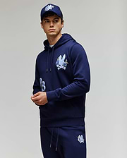 A young man wears the Psycho Bunny MENS AUGUSTE HOODIE - MT0500043, a regular fit navy blue premium cotton hoodie with chenille floral detailing, paired with matching sweatpants and a navy cap, standing against a light gray background.