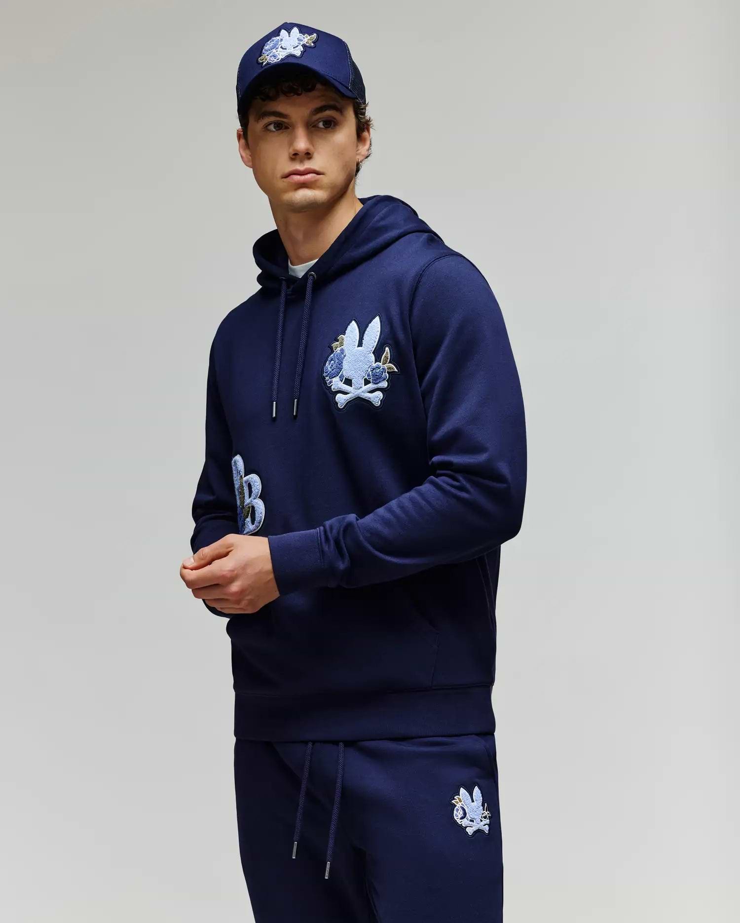 A young man wears the Psycho Bunny MENS AUGUSTE HOODIE - MT0500043, a regular fit navy blue premium cotton hoodie with chenille floral detailing, paired with matching sweatpants and a navy cap, standing against a light gray background.