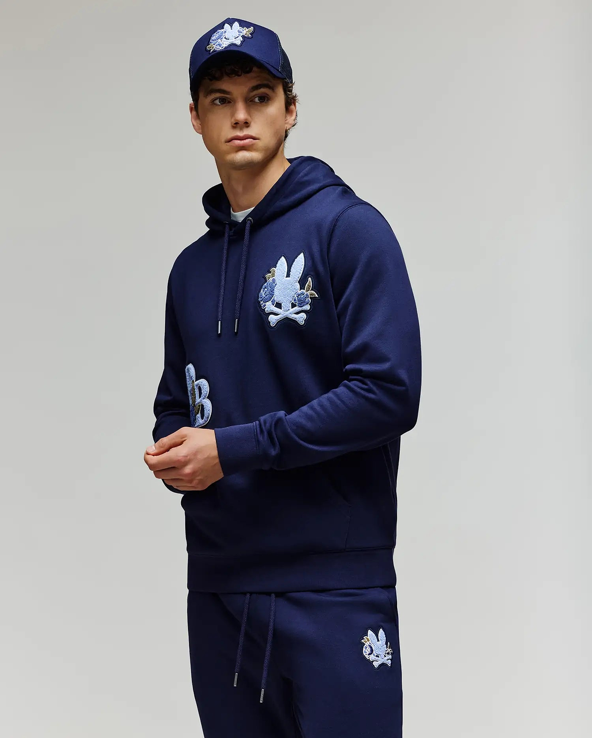 A young man wears the Psycho Bunny MENS AUGUSTE HOODIE - MT0500043, a regular fit navy blue premium cotton hoodie with chenille floral detailing, paired with matching sweatpants and a navy cap, standing against a light gray background.