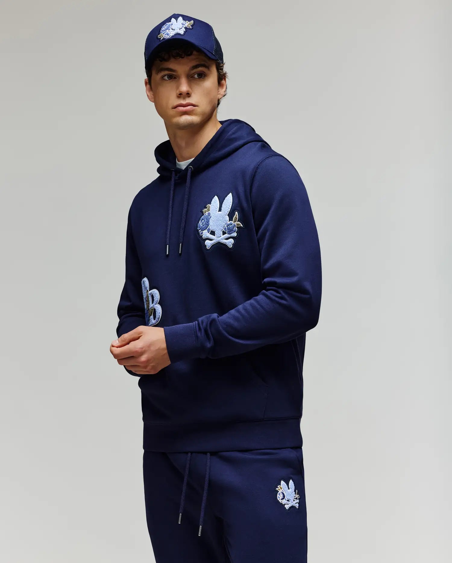 A young man wears the Psycho Bunny MENS AUGUSTE HOODIE - MT0500043, a regular fit navy blue premium cotton hoodie with chenille floral detailing, paired with matching sweatpants and a navy cap, standing against a light gray background.