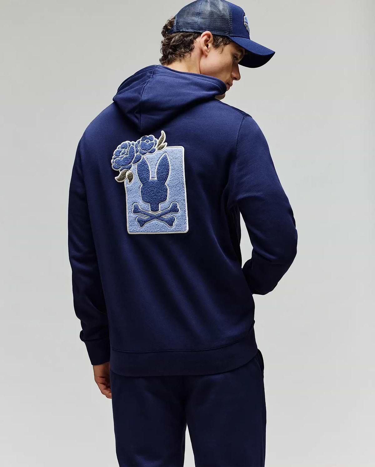 A person wearing a navy blue Psycho Bunny MENS AUGUSTE HOODIE (MT0500043) and cap, with their back turned. The regular fit hoodie features a large rabbit skull and crossbones patch with chenille floral details on the back.
