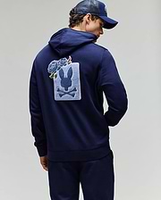 A person wearing a navy blue Psycho Bunny MENS AUGUSTE HOODIE (MT0500043) and cap, with their back turned. The regular fit hoodie features a large rabbit skull and crossbones patch with chenille floral details on the back.