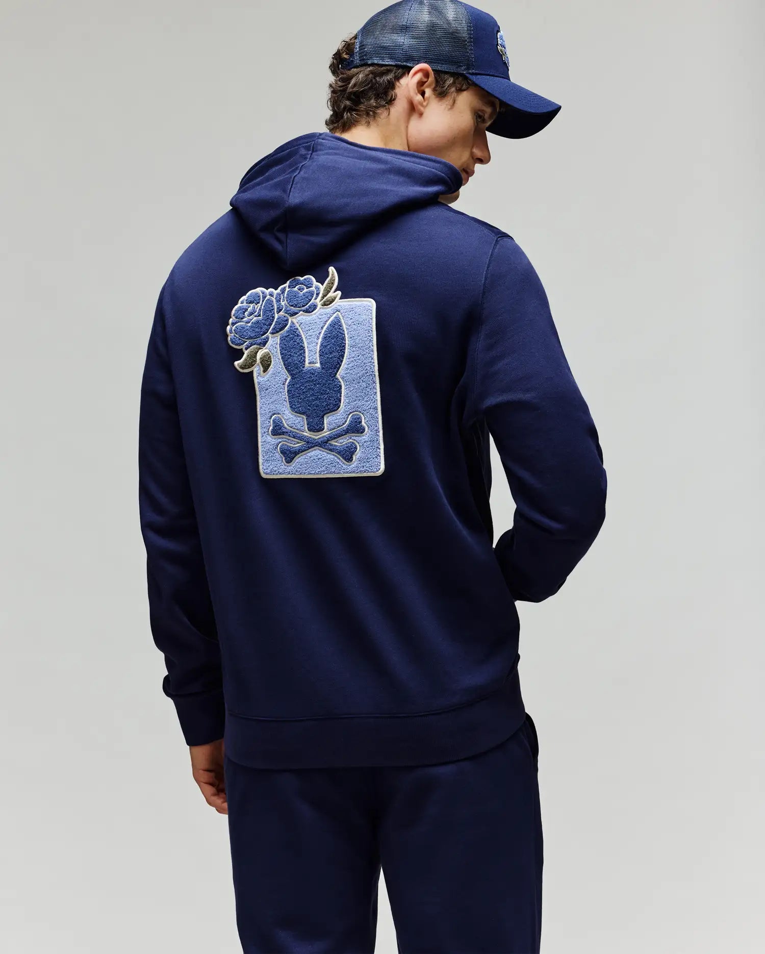 A person wearing a navy blue Psycho Bunny MENS AUGUSTE HOODIE (MT0500043) and cap, with their back turned. The regular fit hoodie features a large rabbit skull and crossbones patch with chenille floral details on the back.