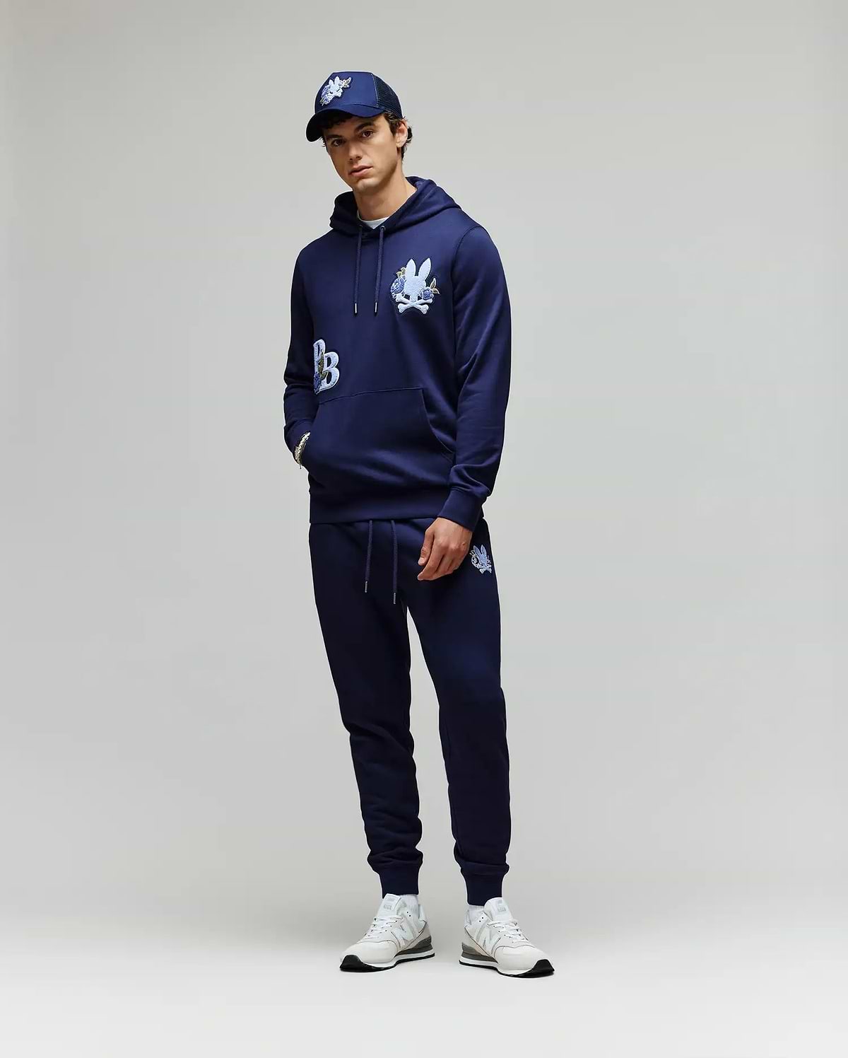 A person stands against a plain background wearing the Psycho Bunny MENS AUGUSTE HOODIE - MT0500043 and matching joggers, both in navy with white graphics, styled with a navy cap and white sneakers. Hands are casually tucked in the pockets.