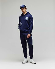A person stands against a plain background wearing the Psycho Bunny MENS AUGUSTE HOODIE - MT0500043 and matching joggers, both in navy with white graphics, styled with a navy cap and white sneakers. Hands are casually tucked in the pockets.