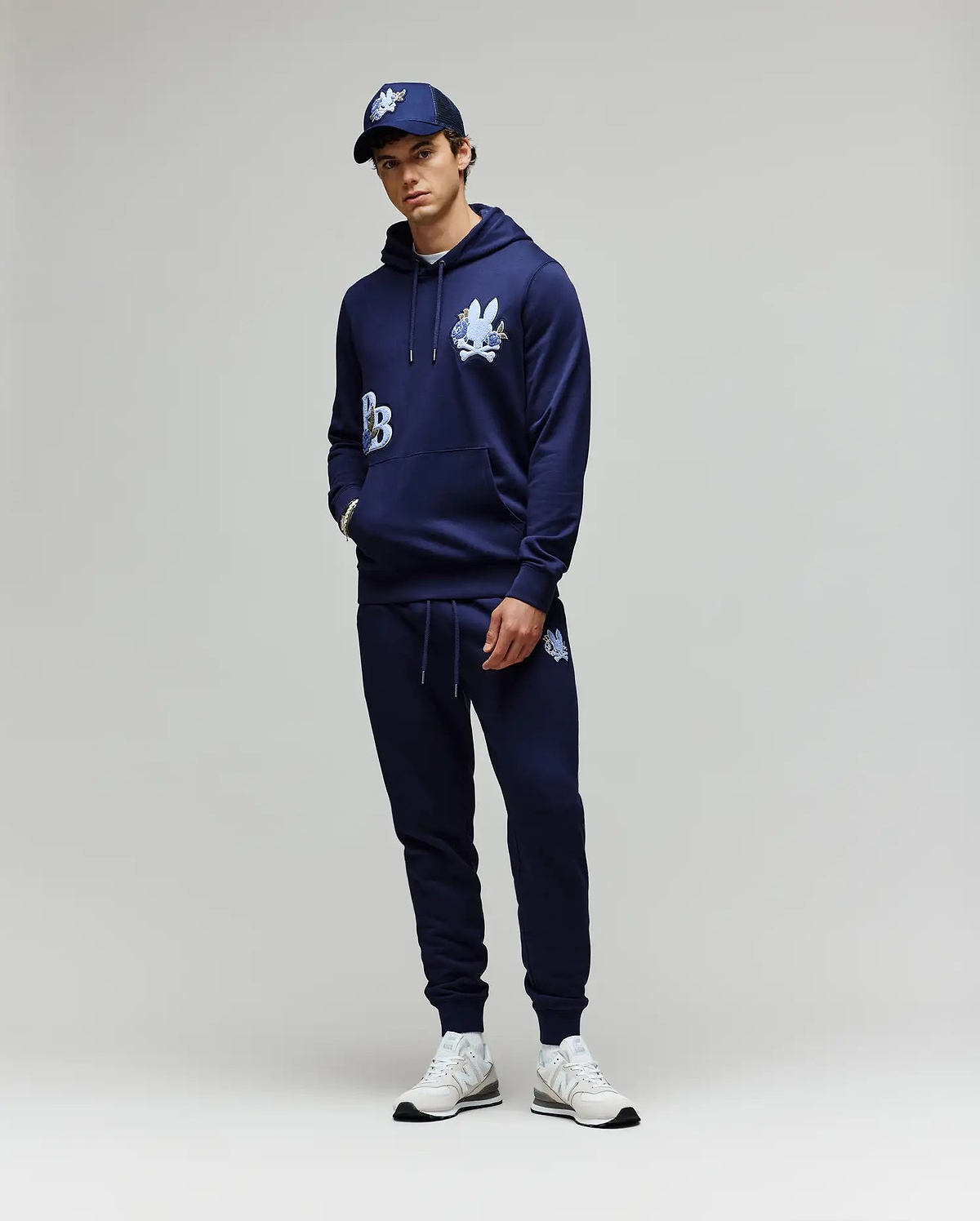 A person stands against a plain background wearing the Psycho Bunny MENS AUGUSTE HOODIE - MT0500043 and matching joggers, both in navy with white graphics, styled with a navy cap and white sneakers. Hands are casually tucked in the pockets.