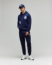 A person stands against a plain background wearing the Psycho Bunny MENS AUGUSTE HOODIE - MT0500043 and matching joggers, both in navy with white graphics, styled with a navy cap and white sneakers. Hands are casually tucked in the pockets.