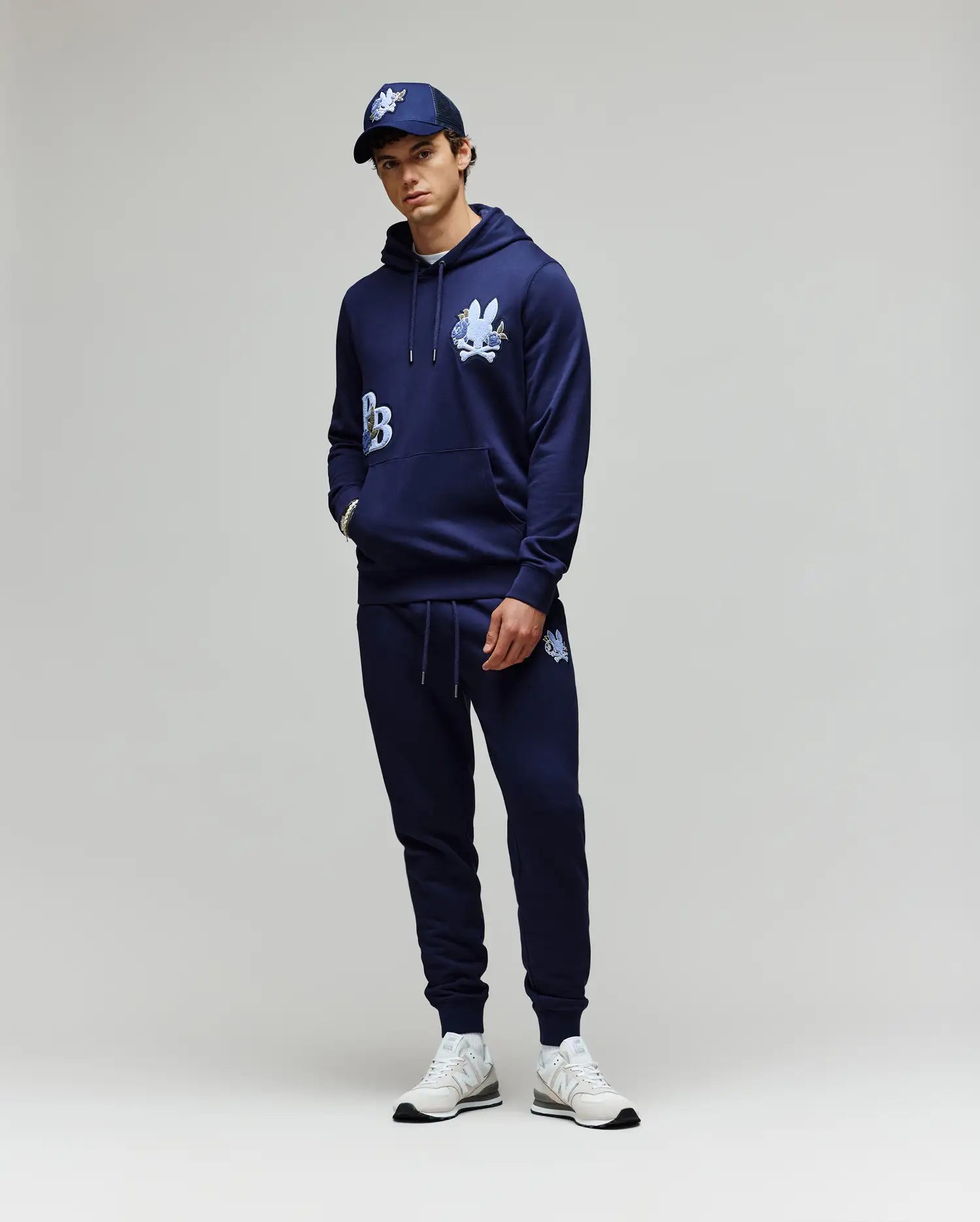 A person stands against a plain background wearing the Psycho Bunny MENS AUGUSTE HOODIE - MT0500043 and matching joggers, both in navy with white graphics, styled with a navy cap and white sneakers. Hands are casually tucked in the pockets.