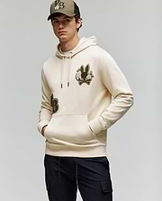 A young man stands with his hands in his pockets, wearing Psycho Bunny's MENS AUGUSTE HOODIE (MT0500043) in beige with chenille floral detailing, black pants, and a matching beige baseball cap against a plain light background.