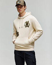 A young man stands with his hands in his pockets, wearing Psycho Bunny's MENS AUGUSTE HOODIE (MT0500043) in beige with chenille floral detailing, black pants, and a matching beige baseball cap against a plain light background.