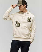 A man wearing the Psycho Bunny MENS AUGUSTE HOODIE - MT0500043 in beige with chenille floral details and a matching cap, looking to the side while adjusting his cap and pairing it with dark pants for a stylish, laid-back vibe.