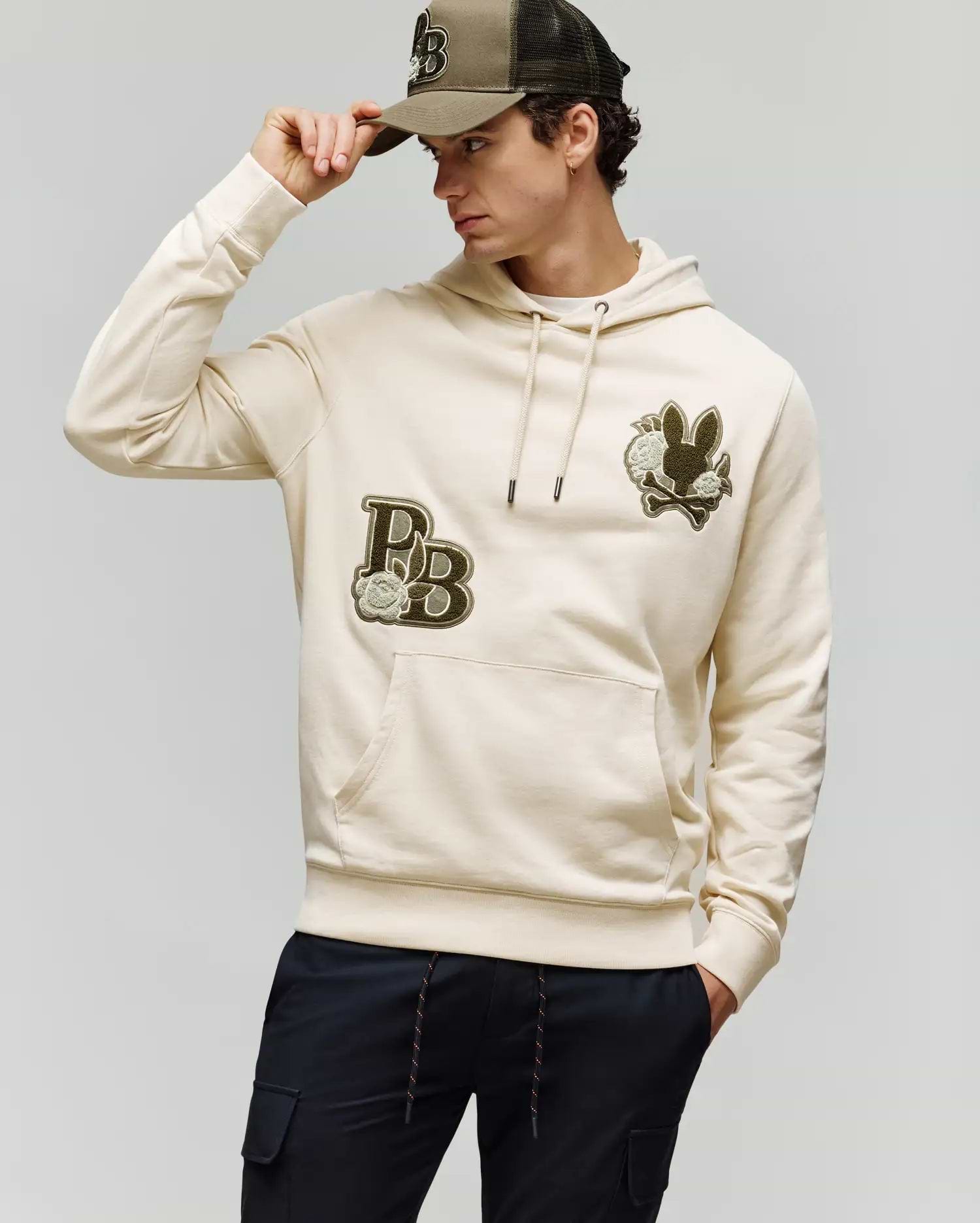 A man wearing the Psycho Bunny MENS AUGUSTE HOODIE - MT0500043 in beige with chenille floral details and a matching cap, looking to the side while adjusting his cap and pairing it with dark pants for a stylish, laid-back vibe.