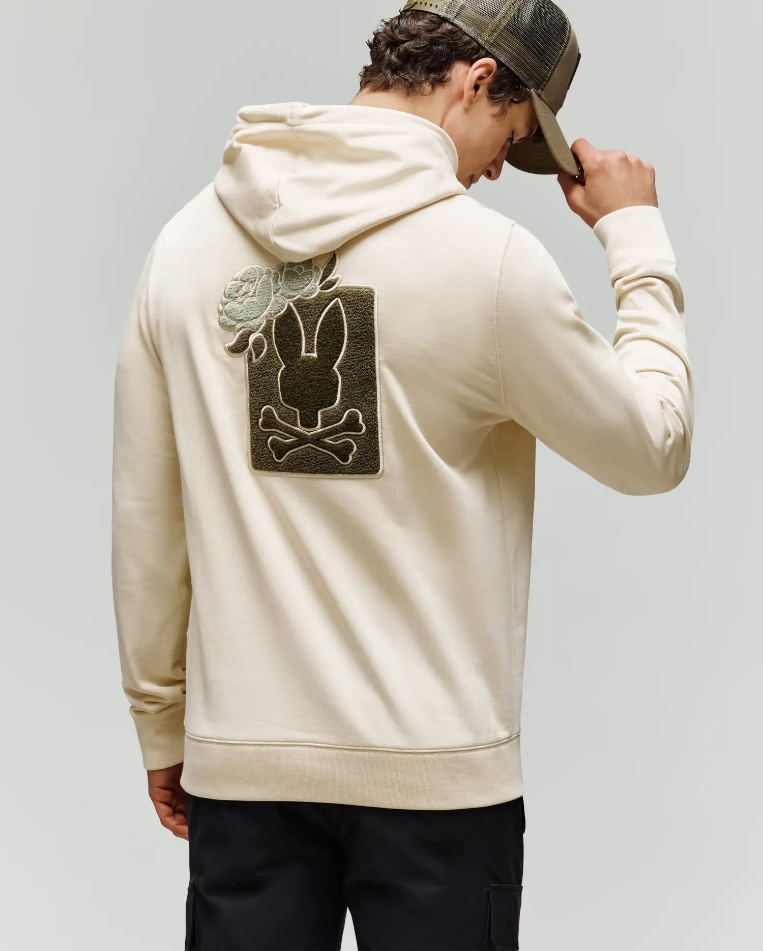 A man in Psycho Bunny’s MENS AUGUSTE HOODIE - MT0500043, a regular-fit premium cotton style, faces away tipping a cap. The back features chenille floral, bunny skull and crossbones, and a rose patch design.