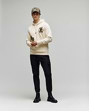A young man stands against a plain background wearing the Psycho Bunny MENS AUGUSTE HOODIE - MT0500043, featuring chenille floral detailing, paired with a matching beige cap, black cargo pants, and black boots.