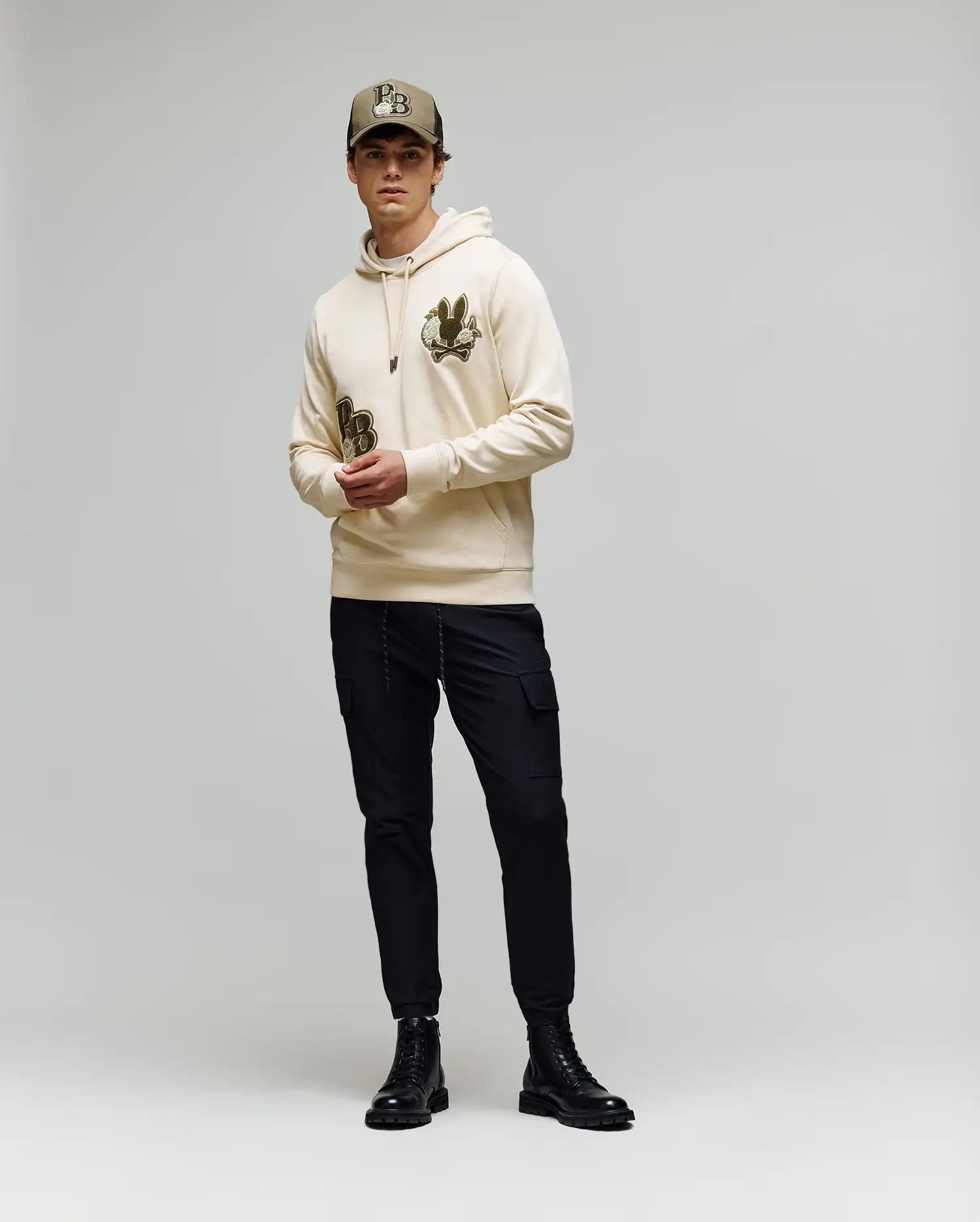 A young man stands against a plain background wearing the Psycho Bunny MENS AUGUSTE HOODIE - MT0500043, featuring chenille floral detailing, paired with a matching beige cap, black cargo pants, and black boots.