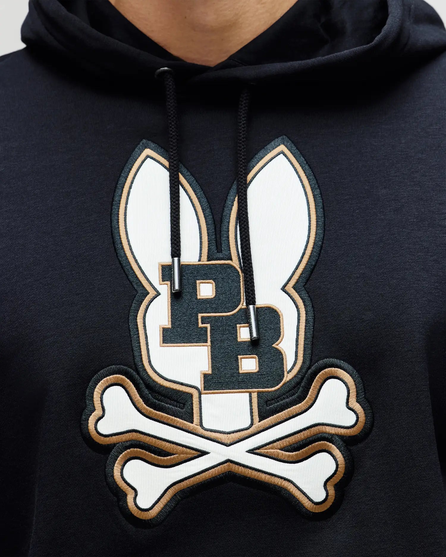 A close-up of the Psycho Bunny MENS WADE CORD PATCH HOODIE (MT0500112), a black premium cotton-blend hoodie featuring an embroidered logo with two bunny ears, bold "PB" initials, and crossed bones underneath.