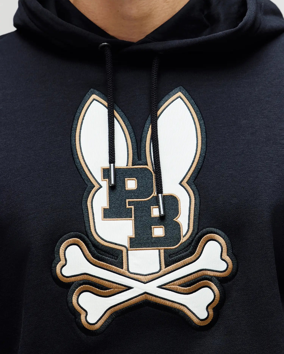 A close-up of the Psycho Bunny MENS WADE CORD PATCH HOODIE (MT0500112), a black premium cotton-blend hoodie featuring an embroidered logo with two bunny ears, bold "PB" initials, and crossed bones underneath.