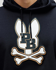 A close-up of the Psycho Bunny MENS WADE CORD PATCH HOODIE (MT0500112), a black premium cotton-blend hoodie featuring an embroidered logo with two bunny ears, bold "PB" initials, and crossed bones underneath.