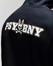 A man wears the Psycho Bunny MENS WADE CORD PATCH HOODIE (MT0500112), crafted from a premium cotton blend and featuring "PSY BNY" plus a skull-and-crossbones with rabbit ears on the back.