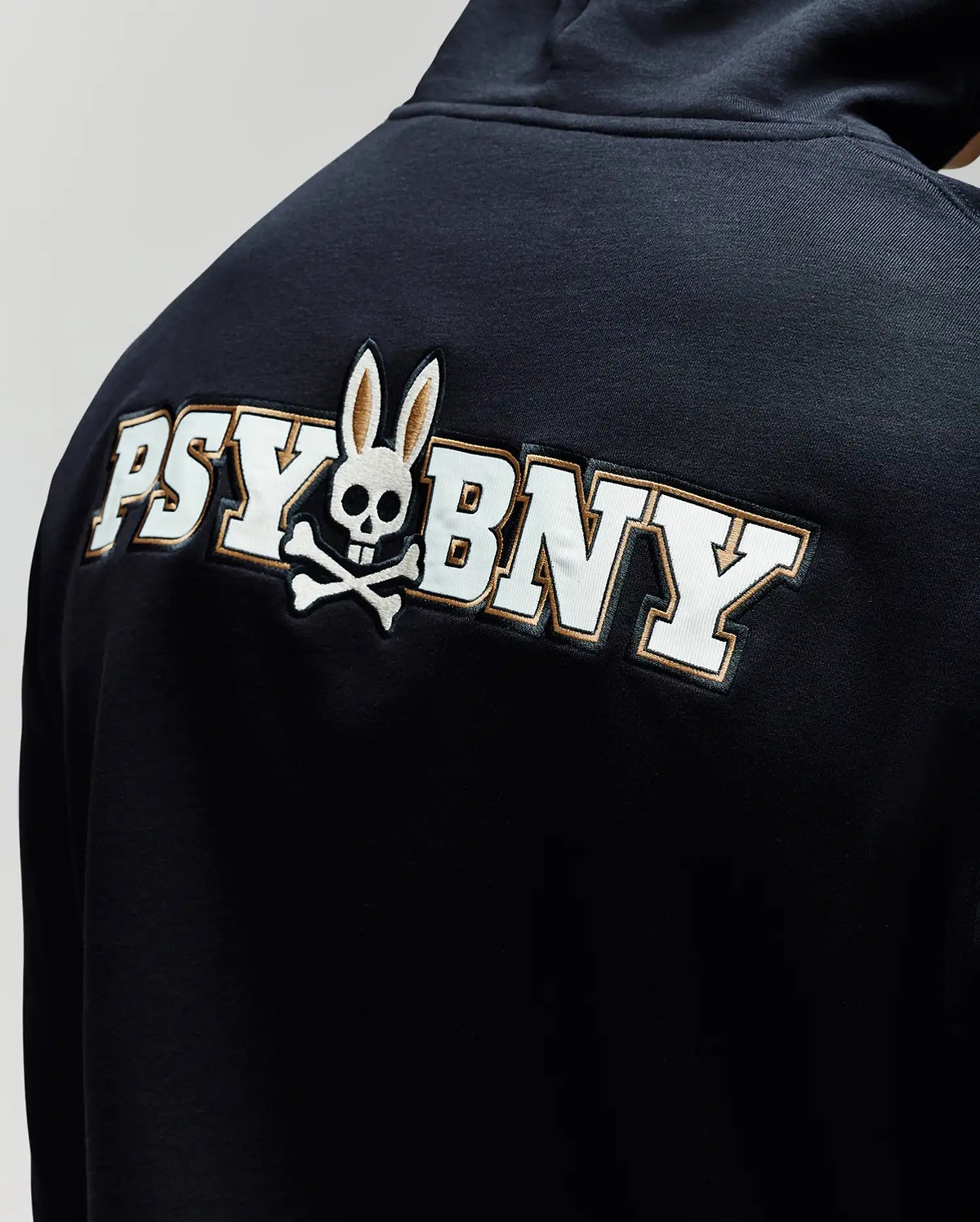 A man wears the Psycho Bunny MENS WADE CORD PATCH HOODIE (MT0500112), crafted from a premium cotton blend and featuring "PSY BNY" plus a skull-and-crossbones with rabbit ears on the back.
