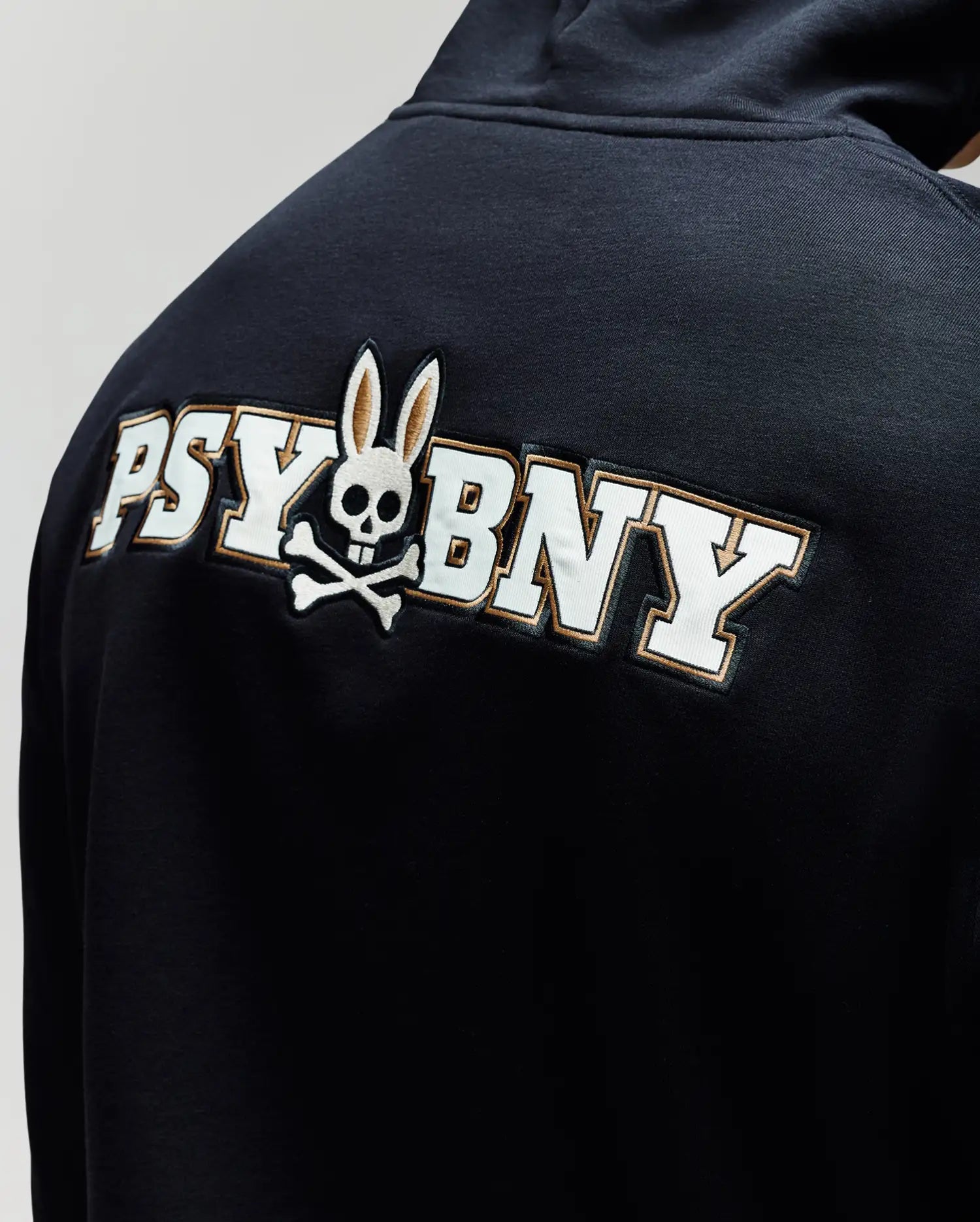A man wears the Psycho Bunny MENS WADE CORD PATCH HOODIE (MT0500112), crafted from a premium cotton blend and featuring "PSY BNY" plus a skull-and-crossbones with rabbit ears on the back.