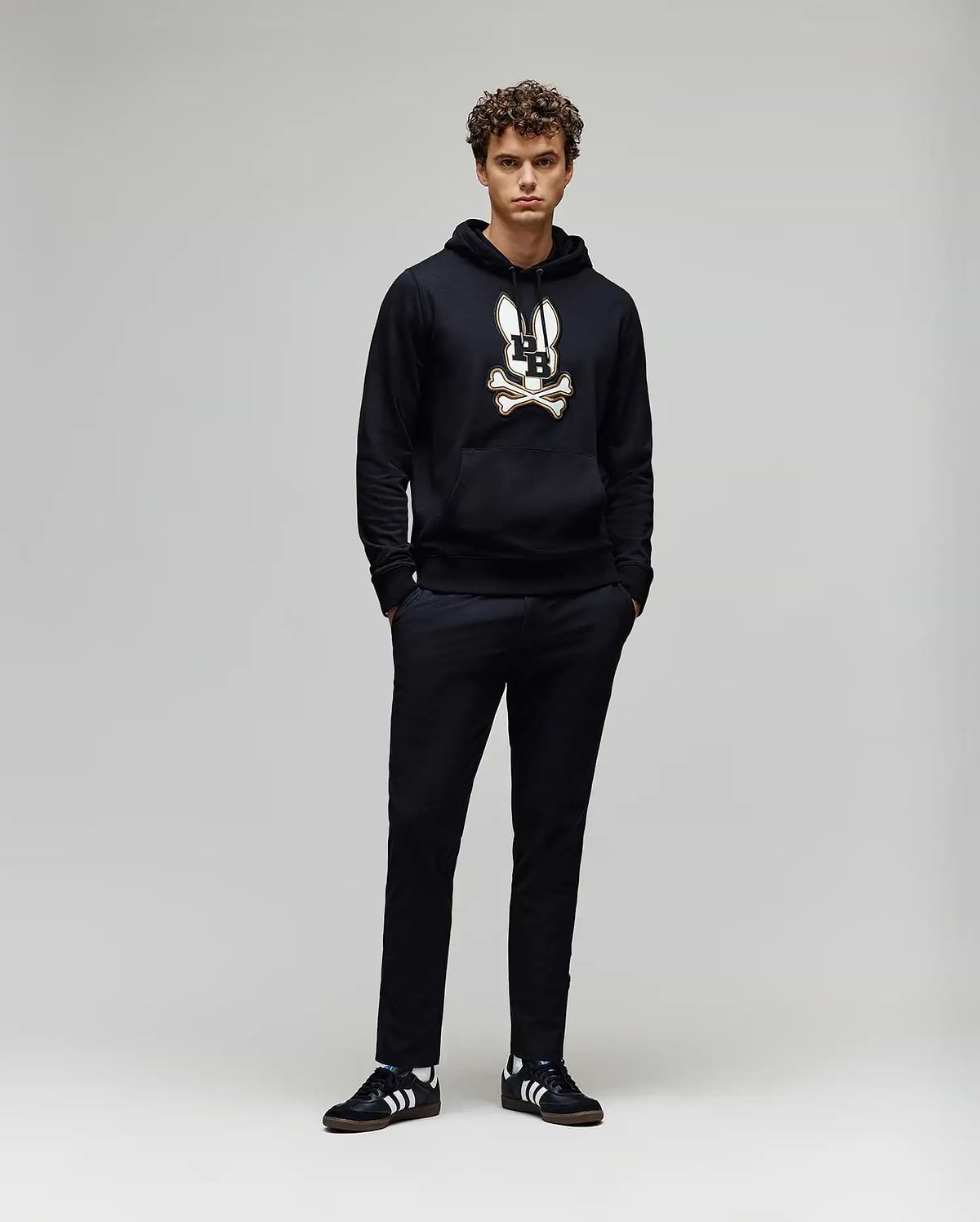 A young man with curly hair stands against a plain background, wearing the Psycho Bunny MENS WADE CORD PATCH HOODIE (MT0500112) in black, paired with black pants and black Adidas sneakers with white stripes. His hands are in his pockets.