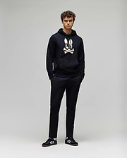 A young man with curly hair stands against a plain background, wearing the Psycho Bunny MENS WADE CORD PATCH HOODIE (MT0500112) in black, paired with black pants and black Adidas sneakers with white stripes. His hands are in his pockets.