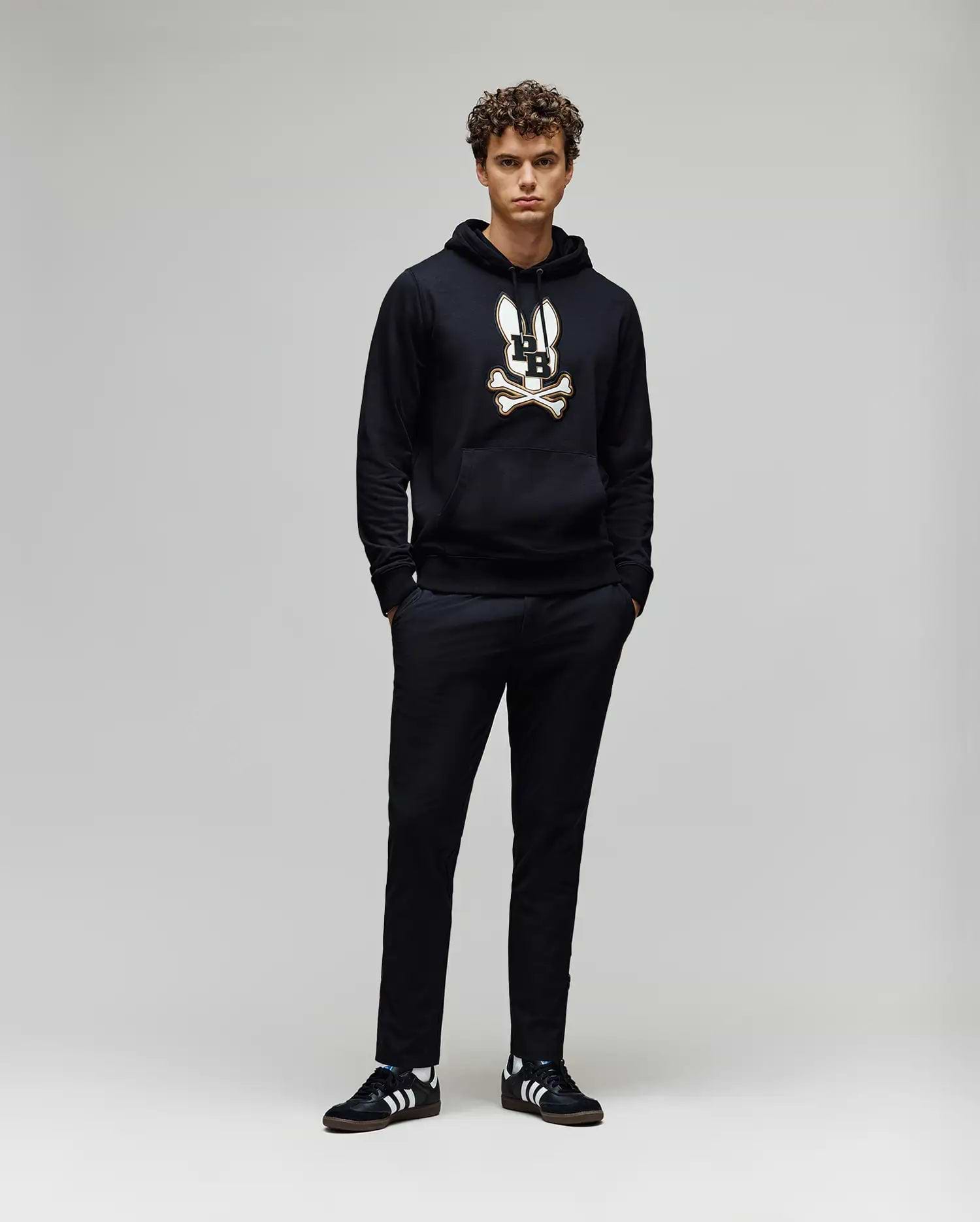 A young man with curly hair stands against a plain background, wearing the Psycho Bunny MENS WADE CORD PATCH HOODIE (MT0500112) in black, paired with black pants and black Adidas sneakers with white stripes. His hands are in his pockets.
