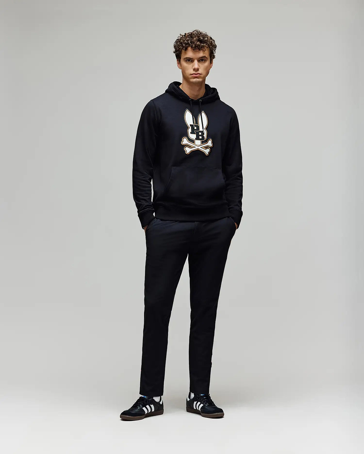 A young man with curly hair stands against a plain background, wearing the Psycho Bunny MENS WADE CORD PATCH HOODIE (MT0500112) in black, paired with black pants and black Adidas sneakers with white stripes. His hands are in his pockets.