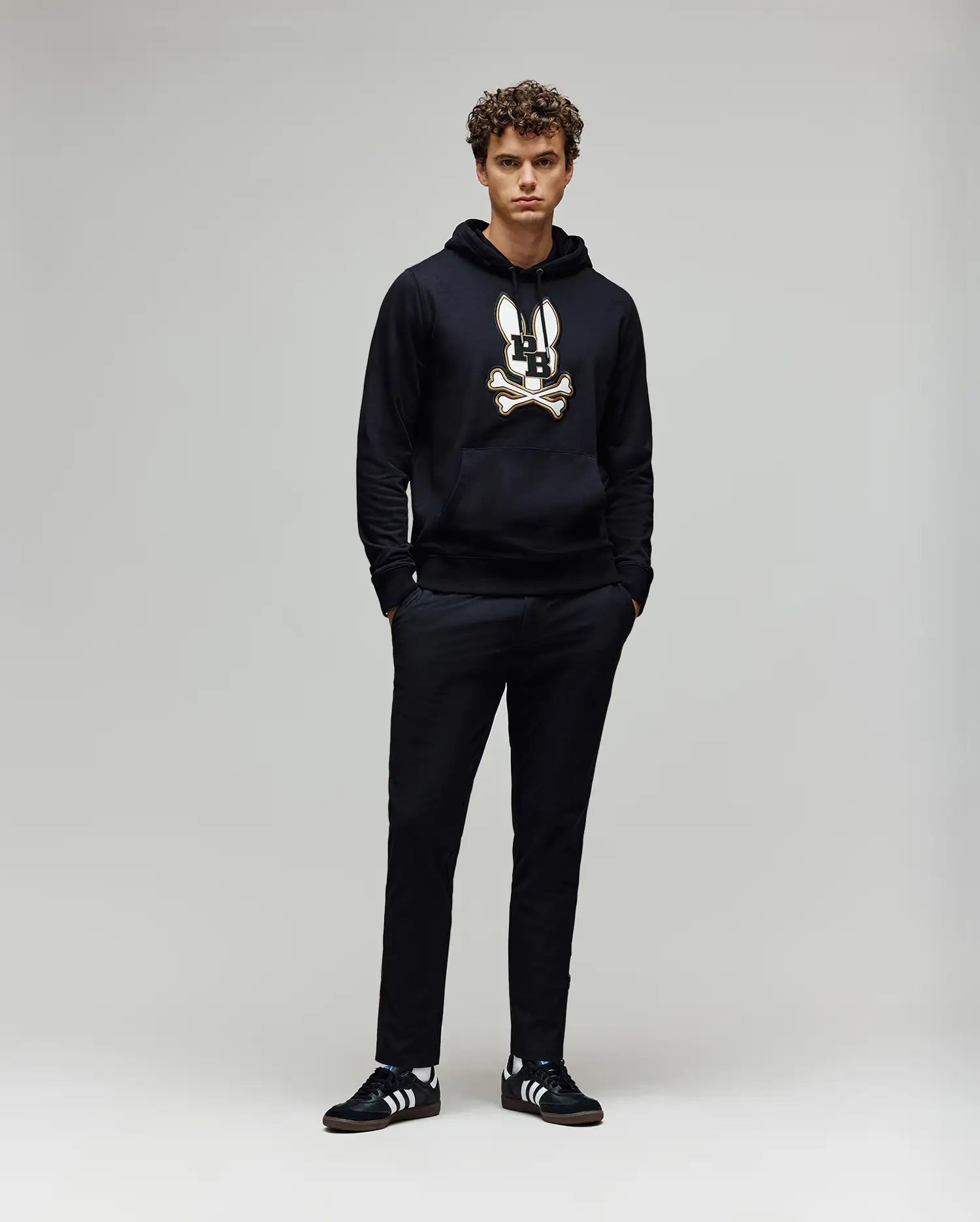 A young man with curly hair stands against a plain background, wearing the Psycho Bunny MENS WADE CORD PATCH HOODIE (MT0500112) in black, paired with black pants and black Adidas sneakers with white stripes. His hands are in his pockets.