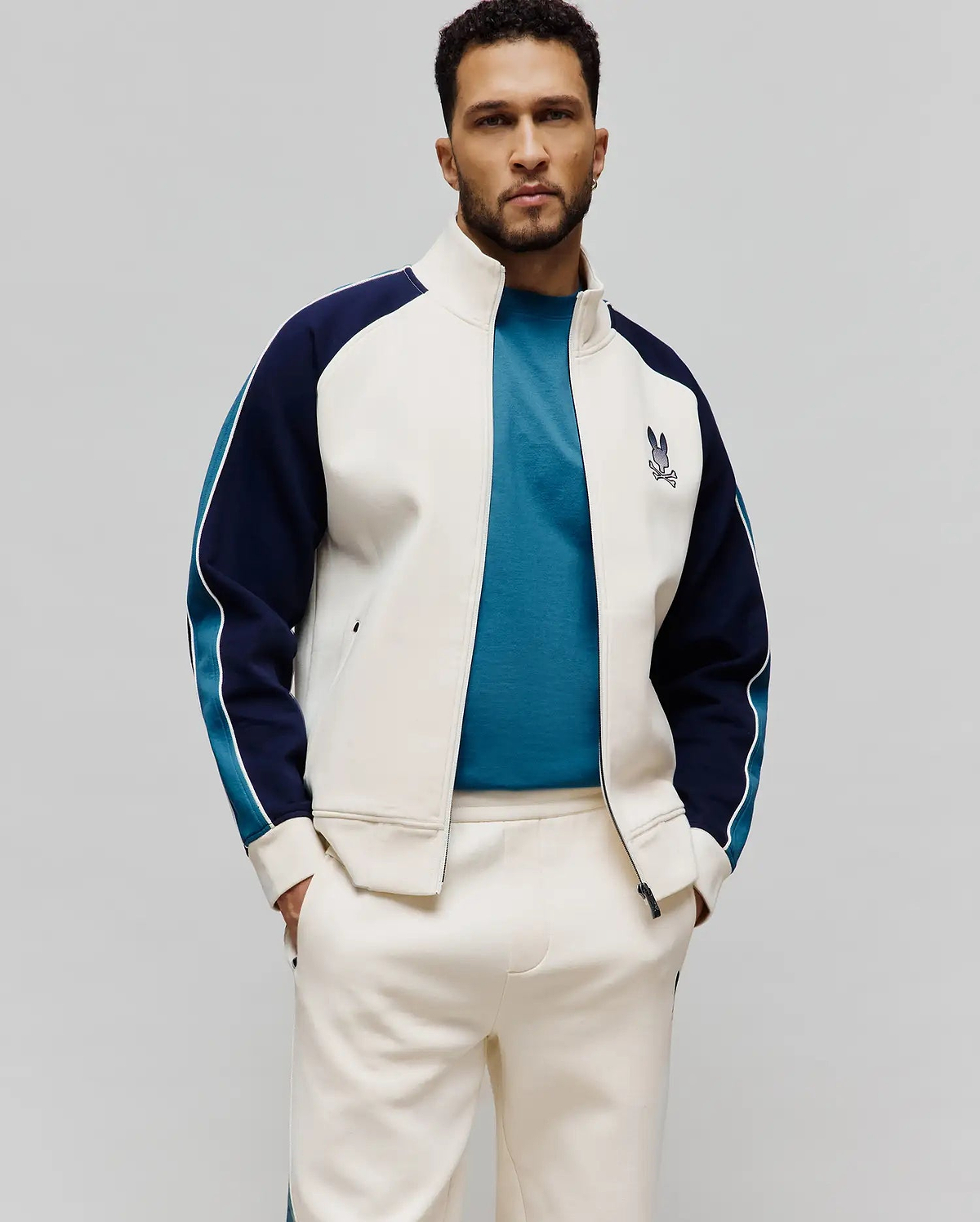 Cream Psycho Bunny smooth knit track jacket with navy and teal color-block sleeves, white piping, an embroidered chest bunny skull logo, a teal t-shirt, and matching cream sweatpants.