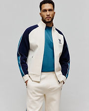 Cream Psycho Bunny smooth knit track jacket with navy and teal color-block sleeves, white piping, an embroidered chest bunny skull logo, a teal t-shirt, and matching cream sweatpants.