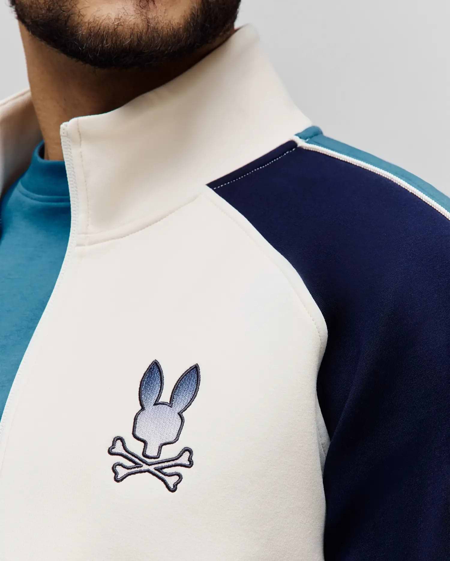 Premium cream smooth knit full-zip track jacket with navy and light blue color block panels, a stand-up collar, and an embroidered Psycho Bunny skull logo on the left chest.
