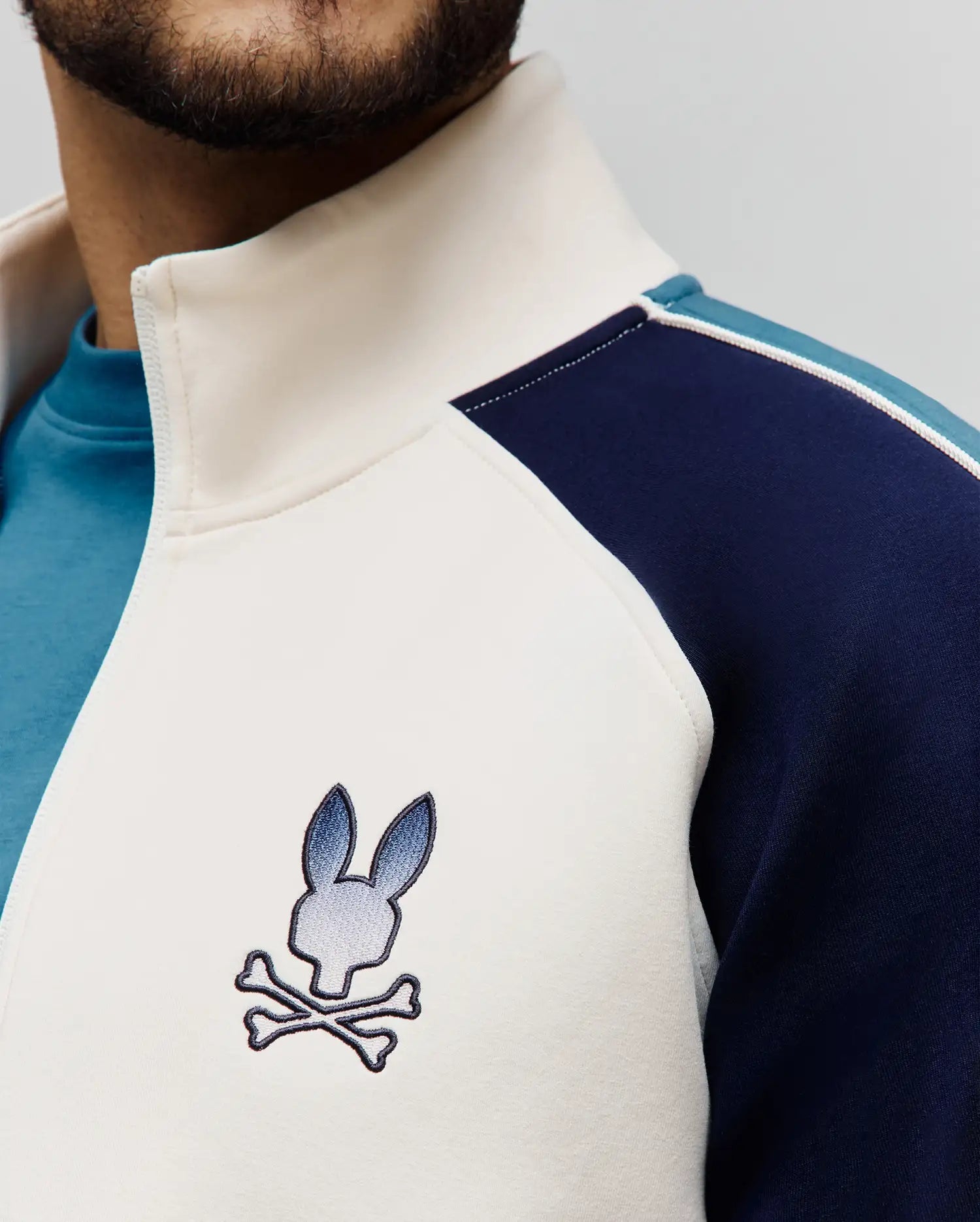 Premium cream smooth knit full-zip track jacket with navy and light blue color block panels, a stand-up collar, and an embroidered Psycho Bunny skull logo on the left chest.