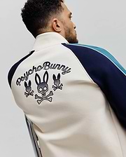 Cream full-zip track jacket with navy and light blue striped raglan sleeves, separated by white piping. Features large embroidered Psycho Bunny script and three bunny skull logos on the back. Classic fit, smooth knit fabric.