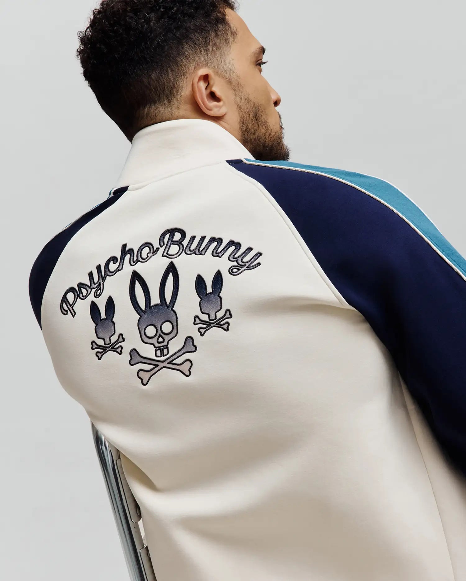 Cream full-zip track jacket with navy and light blue striped raglan sleeves, separated by white piping. Features large embroidered Psycho Bunny script and three bunny skull logos on the back. Classic fit, smooth knit fabric.