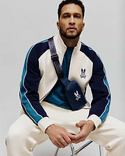 Cream and navy color-block track jacket with teal sleeve stripes, white piping, and an embroidered Psycho Bunny skull logo on chest, paired with cream sweatpants and a navy crossbody bag featuring a pink and white bunny skull.
