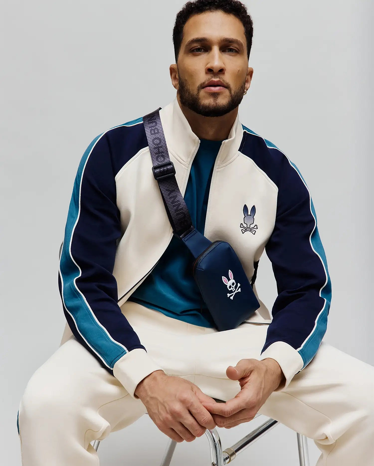 Cream and navy color-block track jacket with teal sleeve stripes, white piping, and an embroidered Psycho Bunny skull logo on chest, paired with cream sweatpants and a navy crossbody bag featuring a pink and white bunny skull.