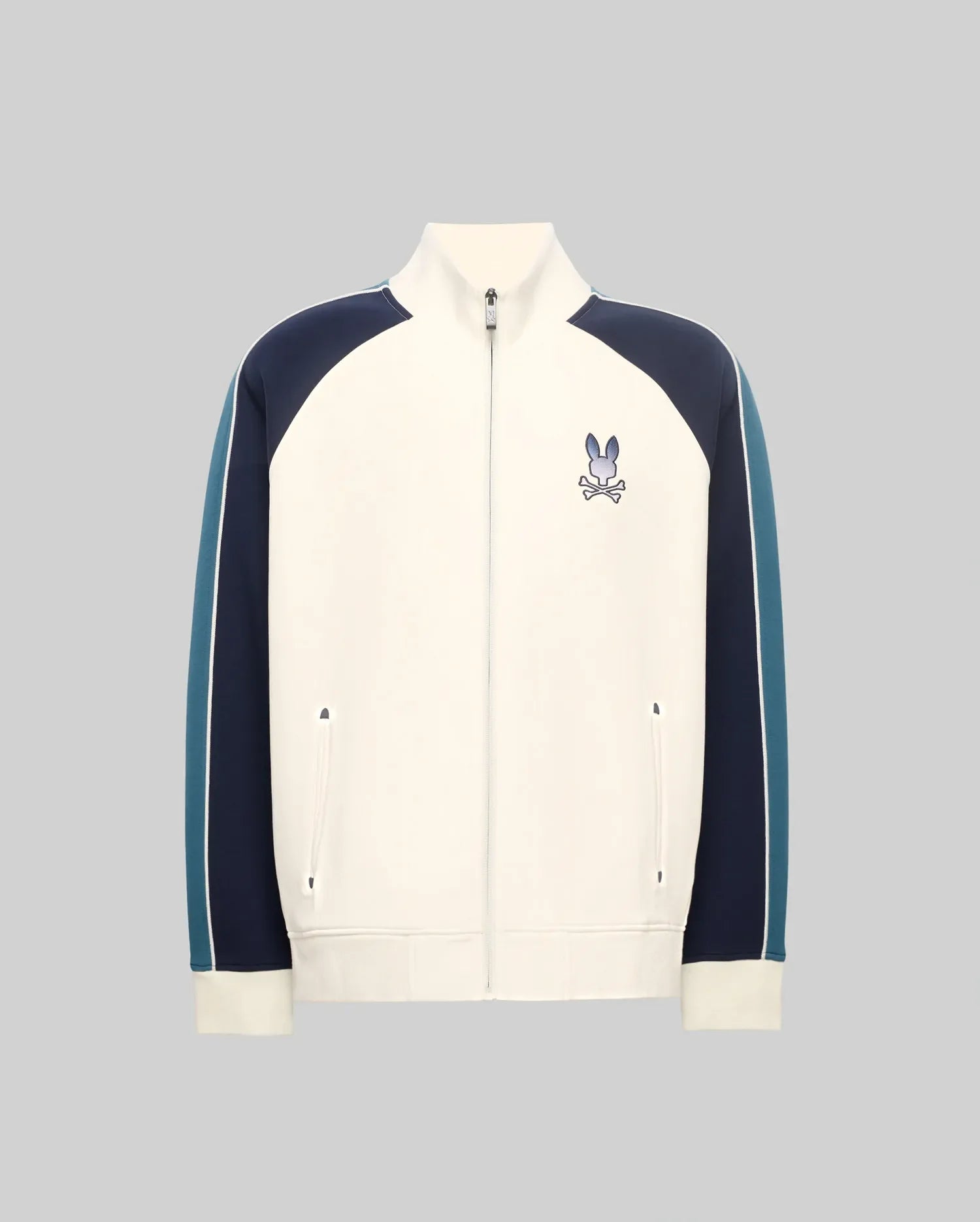 Cream full-zip track jacket with navy and teal color-block sleeves featuring a white stripe, classic fit, smooth knit, stand collar, and an embroidered Psycho Bunny skull logo on the left chest.