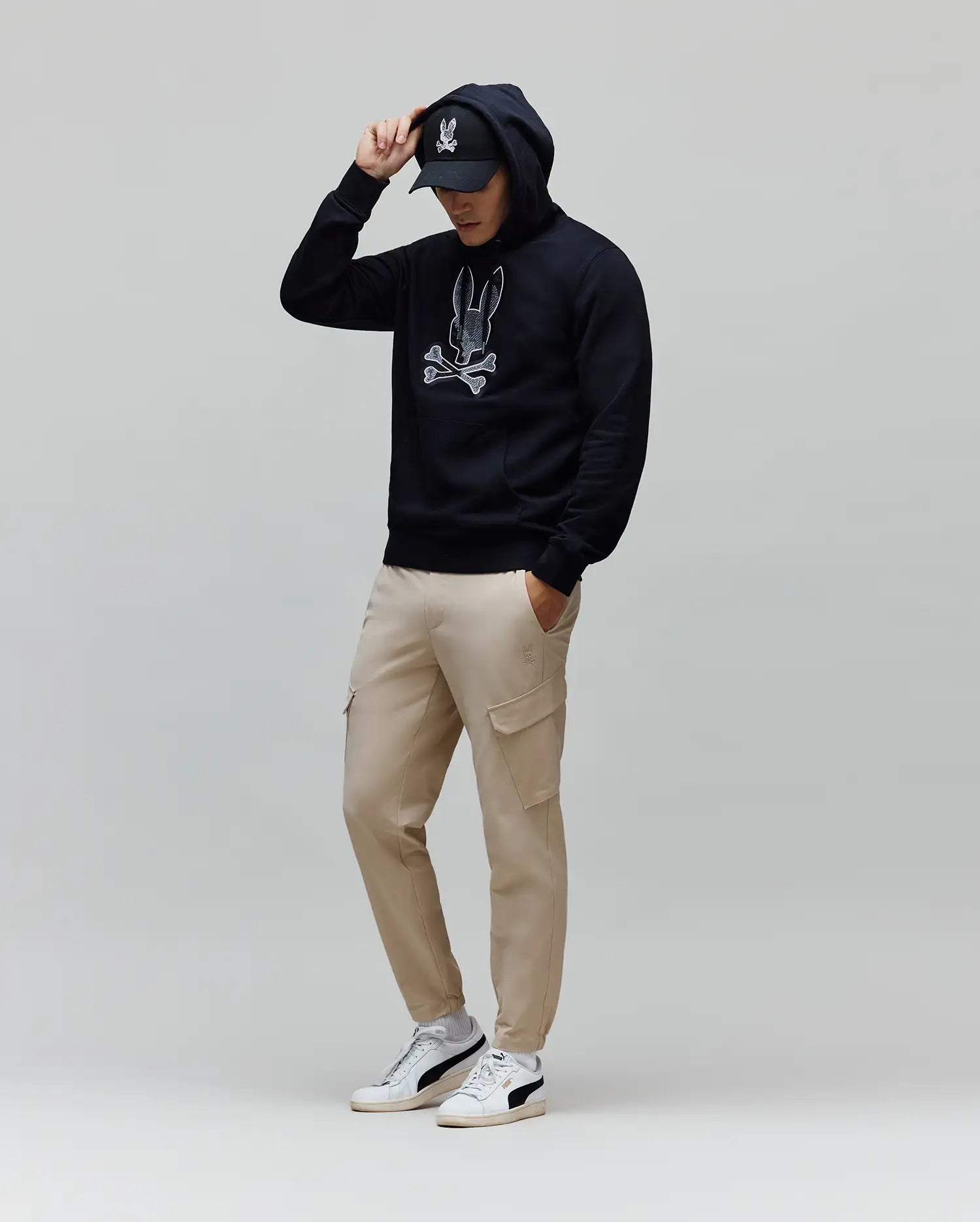 Mens Olivier Embroidered Hoodie: Relaxed fit navy French terry hoodie featuring a large white embroidered Psycho Bunny skull logo on the chest, complemented by beige cargo pants and a black logo cap.